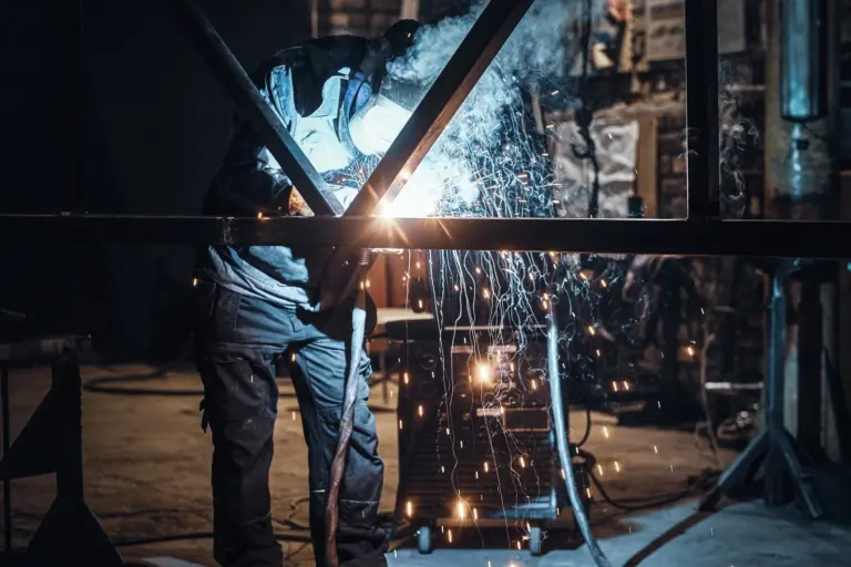 Ein Mann schweißt Stahl in einer Fabrik, umgeben von Maschinen und Werkbänken.