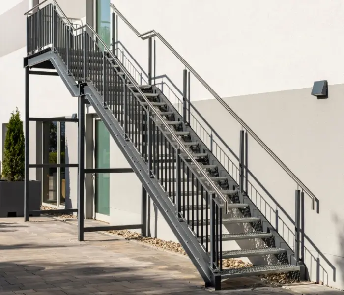 Treppenanlage mit Geländer, die zu einem Gebäude führt, umgeben von Pflanzen und einem klaren Himmel.