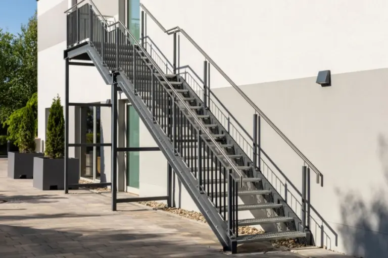 Treppenanlage mit Geländer, die zu einem Gebäude führt, umgeben von Pflanzen und einem klaren Himmel.